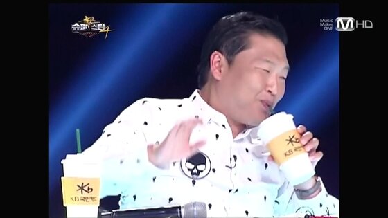 본문 이미지 - 슈스케4에 심사위원으로 출연한 '강남스타일'의 가수 싸이가 'KB국민카드'라고 적힌 컴홀더를 이용해 음료수를 마시고 있다.  © News1  
