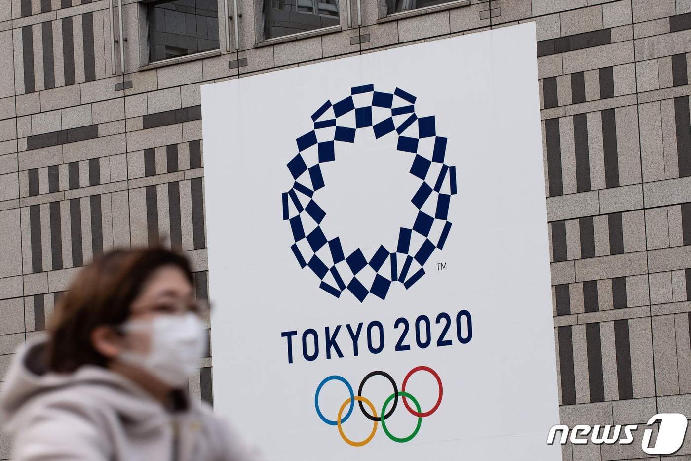 본문 이미지 - 2020년 4월 23일 마스크를 쓴 여성이 도쿄 도청 외벽에 걸린 도쿄 2020 올림픽 게임 로고 앞에서 자전거를 타고 있다. ⓒ AFP=뉴스1