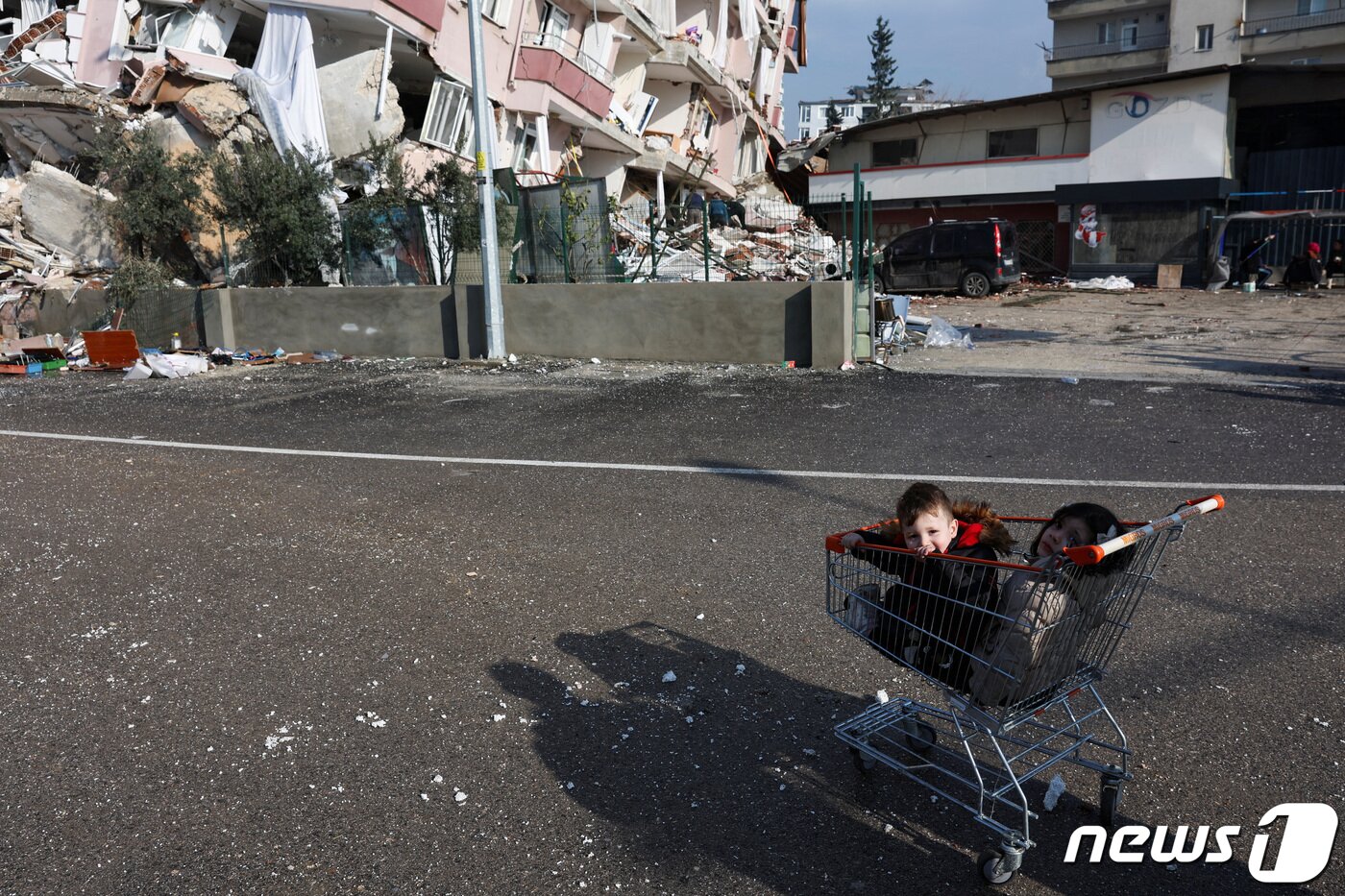 본문 이미지 - 7일(현지시간) 튀르키예 하떠이 한 주택가에 두 꼬마가 시장 카트 속에 쪼그려 앉아 있다. 아이들의 뒤로는 강진으로 무너진 아파트와 잔해가 뒤죽박죽 섞여 있다. ⓒ 로이터=뉴스1 ⓒ News1 권진영 기자