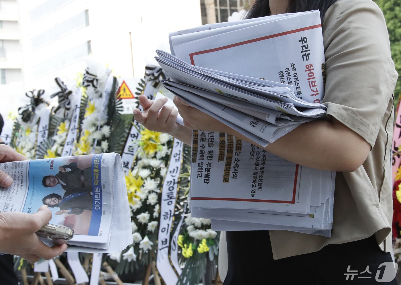 본문 이미지 - 3일 오전 서울 용산구 하이브 사옥 앞에서 방탄소년단(BTS) 한 팬이 성명서가 담긴 신문을 나눠주고 있다. 그 뒤로는 BTS 지지모임이 보낸 근조화환이 세워져 있다. 이들은 하이브에 소속 아티스트 BTS 명예훼손 법적절차 진행을 촉구했다.  ⓒ News1 권현진 기자