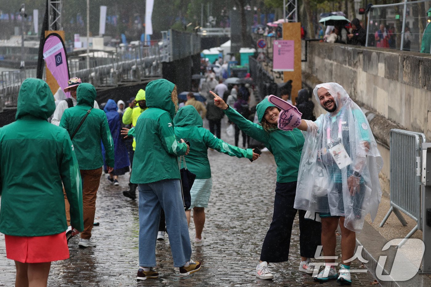 본문 이미지 - 2024 파리올림픽 개회식이 열리는 27일(한국시간) 프랑스 파리 센강에 마련된 관중석 인근에서 자원봉사자들이 비를 맞으며 카메라를 향해 미소를 보이고 있다. (공동취재) 2024.7.27/뉴스1 ⓒ News1 이동해 기자