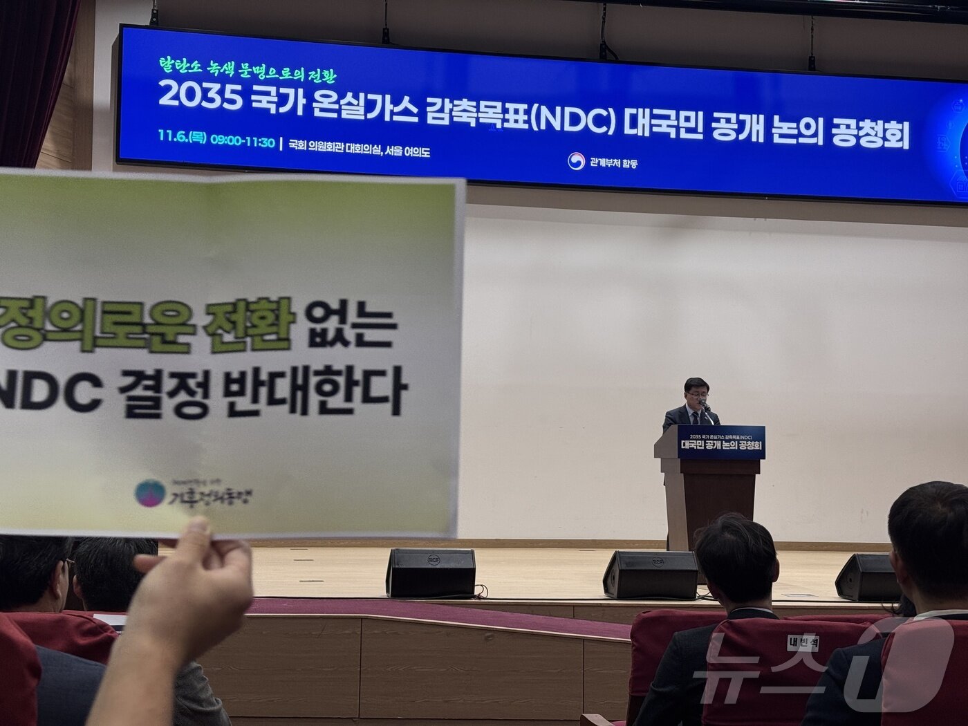 본문 이미지 - 6일 서울 영등포구 국회의원회관 대회의실에서 열린 2035 국가온실가스 감축목표 대국민 공개 논의 공청회에서 김성환 장관이 발언 중인 가운데 환경단체 회원들이 손피켓을 들어보이고 있다. 기후에너지환경부는 2035년 국가온실가스감축목표(NDC)를 2018년 대비 50~60% 감축 범위에서 확정할 예정이다. 2025.11.6/뉴스1 ⓒ News1 황덕현 기후환경전문기자