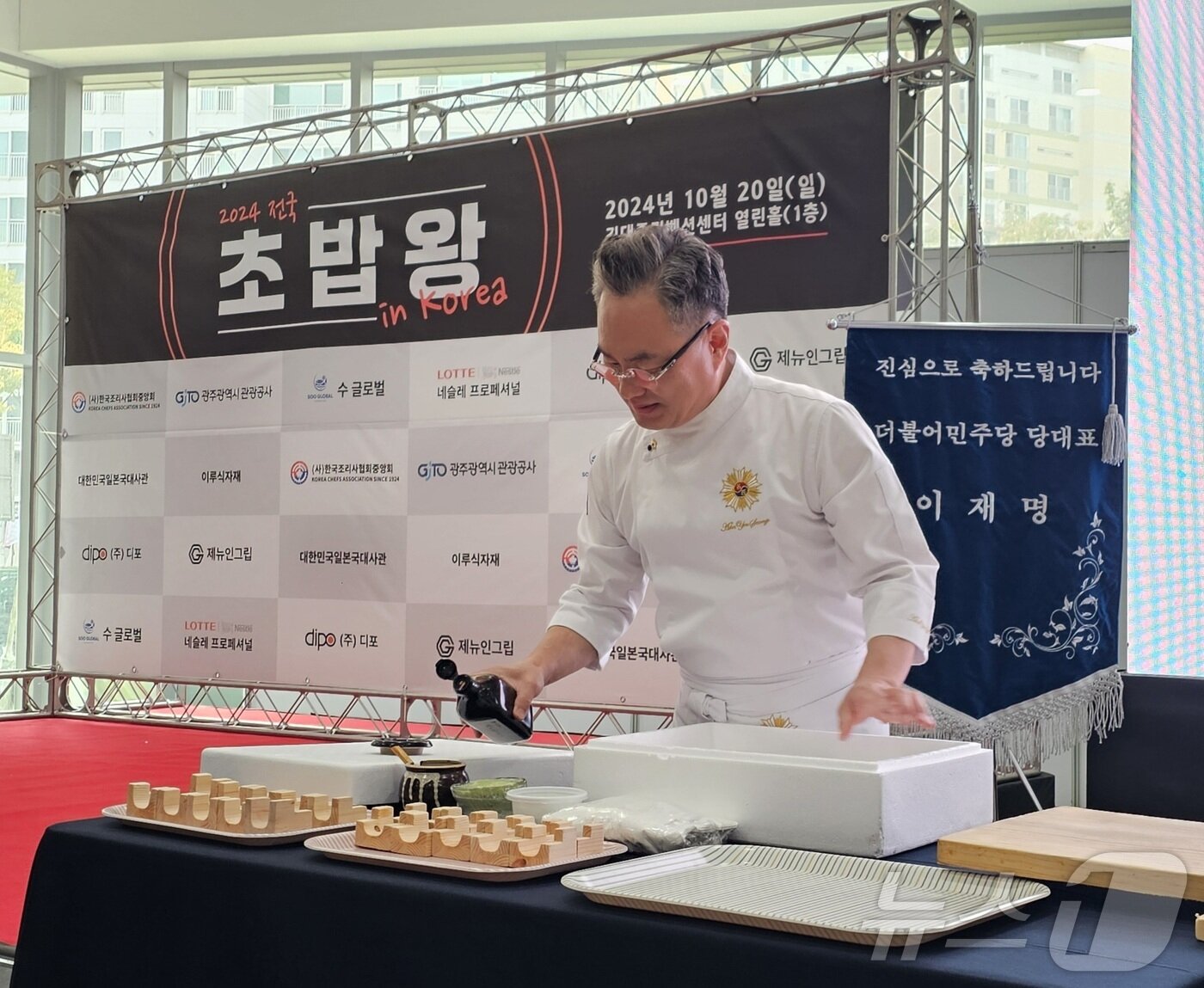 본문 이미지 - 2024년 10월 20일 김대중컨벤션센터에서 ‘전국초밥왕대회’를 개최한 안유성 조리명장이 자연산 다금바리 초밥 시연을 준비하는 뒤로 당시 이재명 더불어민주당 대표의 축하기가 놓였다. 2025.4.25./뉴스1 ⓒ News1 서충섭 기자