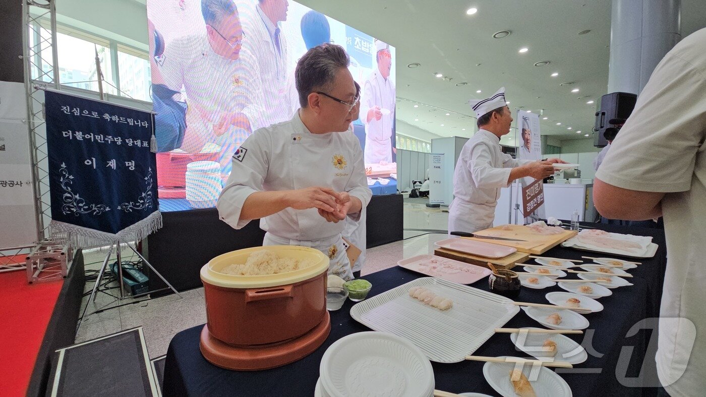 본문 이미지 - 2024년 10월 20일 김대중컨벤션센터에서 ‘전국초밥왕대회’를 개최한 안유성 조리명장이 1㎏당 30만 원에 달하는 자연산 다금바리 초밥 시연을 준비하고 있다. 뒤편엔 이재명 당시 더불어민주당 대표의 축하기가 놓였다. ⓒ News1 서충섭 기자