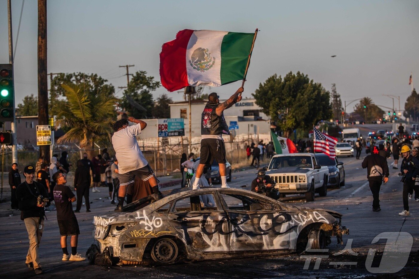본문 이미지 - 7일(현지시간) 미국 LA에서 열린 연방 이민법 집행에 대한 항의 시위에서 시위대들이 불에 탄 차량 위에서 멕시코 국기를 흔들고 있다. ⓒ AFP=뉴스1