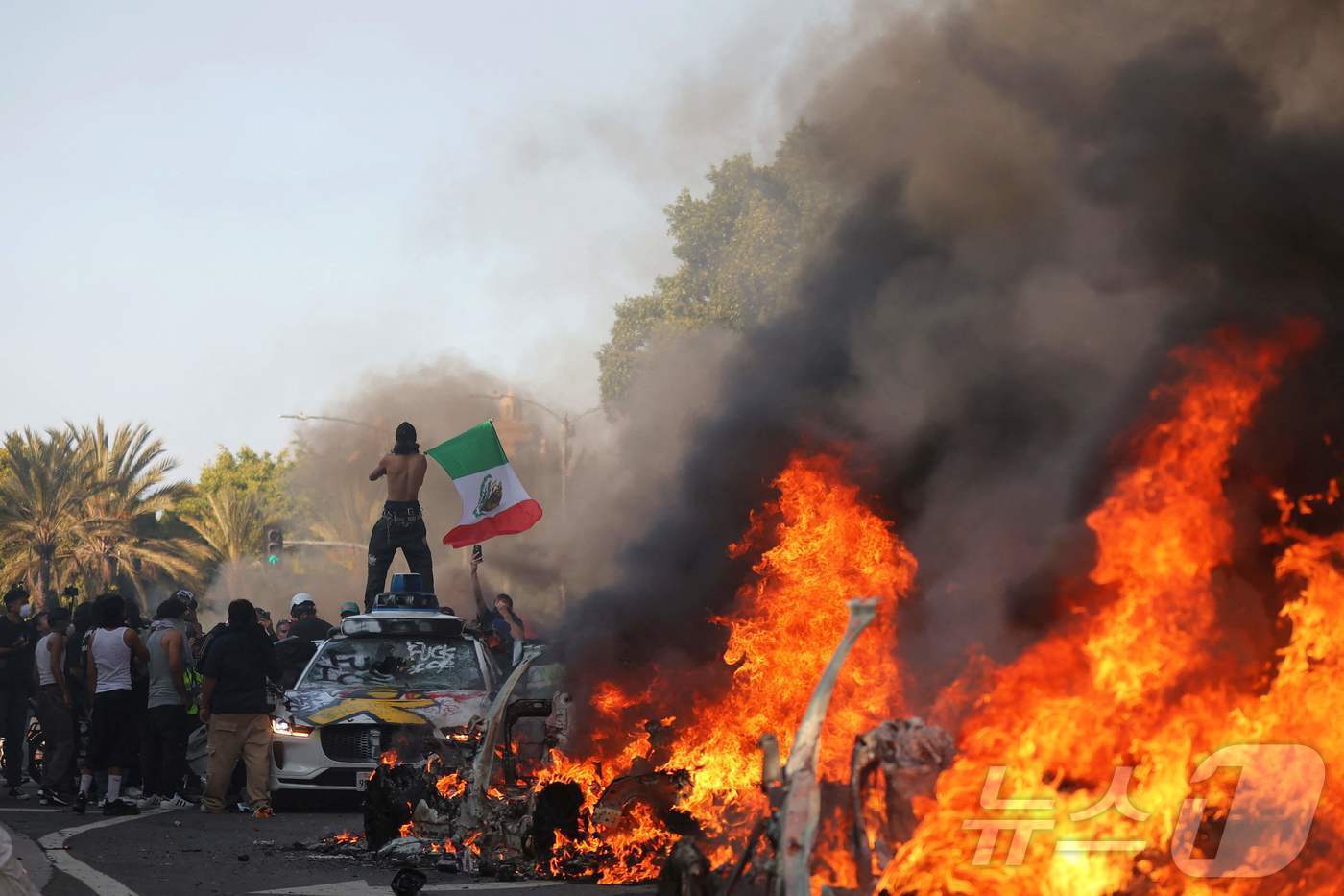 본문 이미지 - 8일(현지시간) 미국 캘리포니아주 LA 시청 인근에서 열린 연방 이민 단속에 항의하는 시위에서 한 남자가 불타는 차량 뒤에서 멕시코 국기를 흔들고 있다. ⓒ AFP=뉴스1