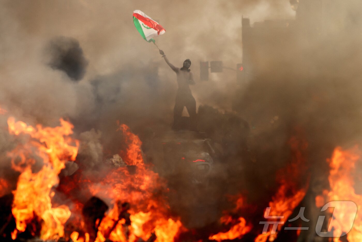 본문 이미지 - 한 남자가 로스앤젤레스 시청 근처에서 연방 이민 단속에 반대하는 시위 중에 연기가 나고 불길이 치솟는 차량 옆에서 멕시코 국기를 흔들고 있다. 2025.6.8 ⓒ 로이터=뉴스1 ⓒ News1 강민경 기자