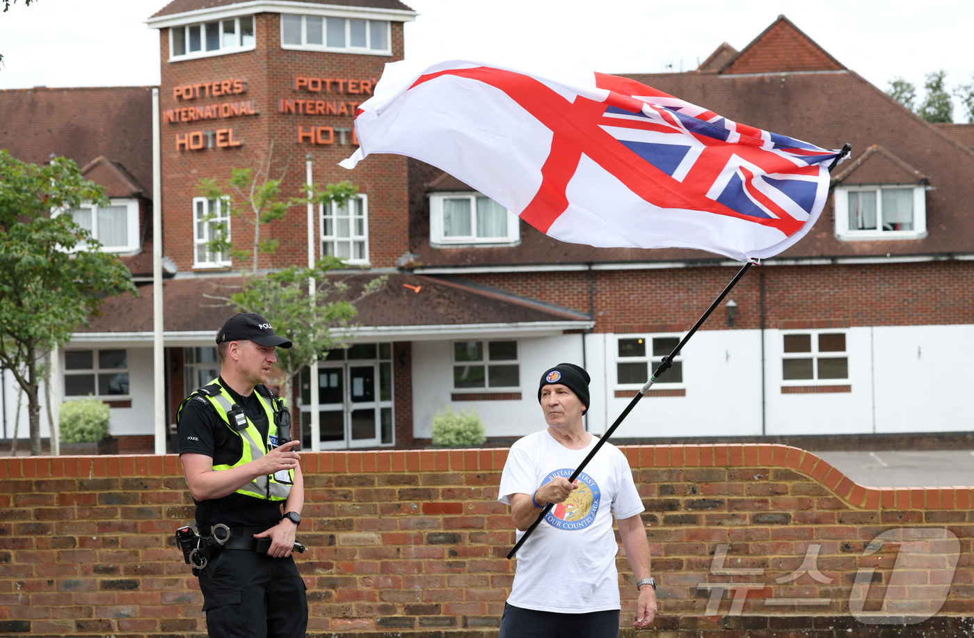 (올더숏 로이터=뉴스1) 김경민 기자 = 2024년 8월 영국 올더숏에서 열린 반이민 시위에서 한 경찰관이 이민자 수용소 호텔 밖에서 영국 해군기를 흔드는 한 남성에게 자리를 비켜 …