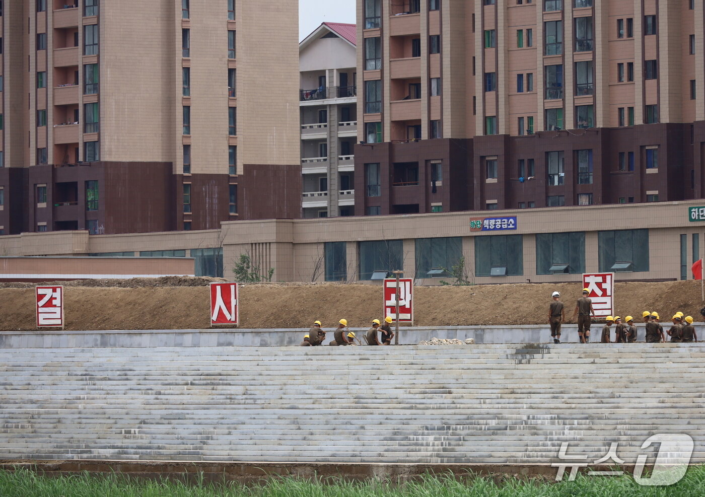 본문 이미지 - 북의 군인건설자들이 새로 건설된 위화도 주택단지 앞에서 제방 공사와 도로정비를 하고 있다. 주택단지 2층에 건설된 식량공급소와 병원 등이 보인다. &#40;평화경제연구소 제공&#41; 
