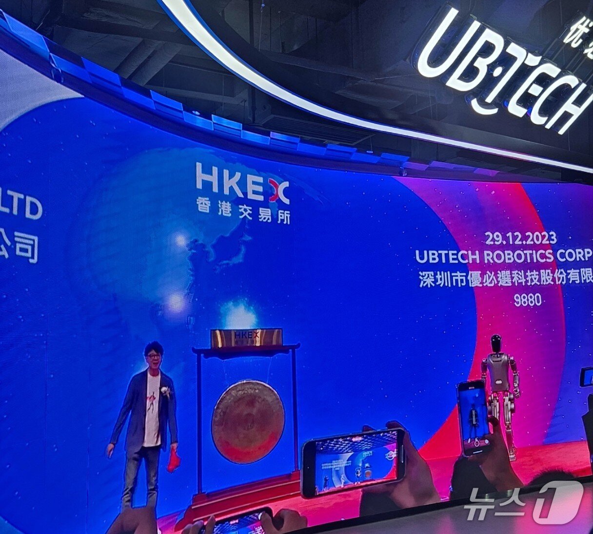 본문 이미지 - 유비테크 창업자 저우젠이 2023년 말 홍콩증권거래소 상장 행사에 자사 휴머노이드 로봇과 함께 징을 치기 위해 준비하고 있는 모습.