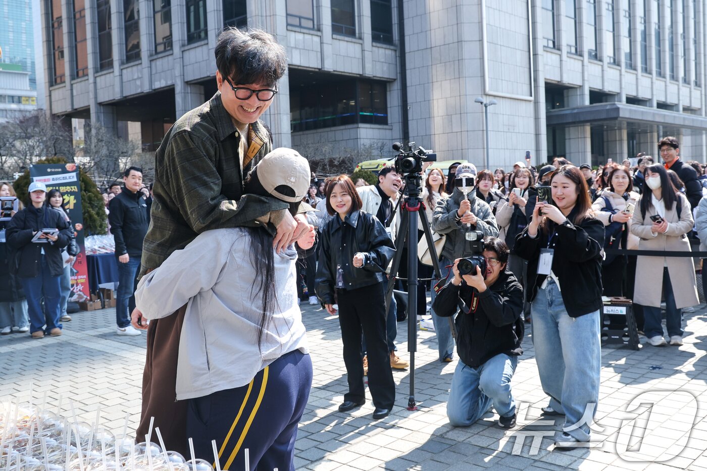 본문 이미지 - 영화 '왕과 사는 남자'의 장항준 감독이 12일 서울 중구 프레스센터 앞에서 1,200만 관객 돌파를 기념해 커피차를 마련, 팬들에게 커피를 나눠주던 중 한 팬이 장 감독을 번쩍 안아들고 있다. 2026.3.12 ⓒ 뉴스1 안은나 기자