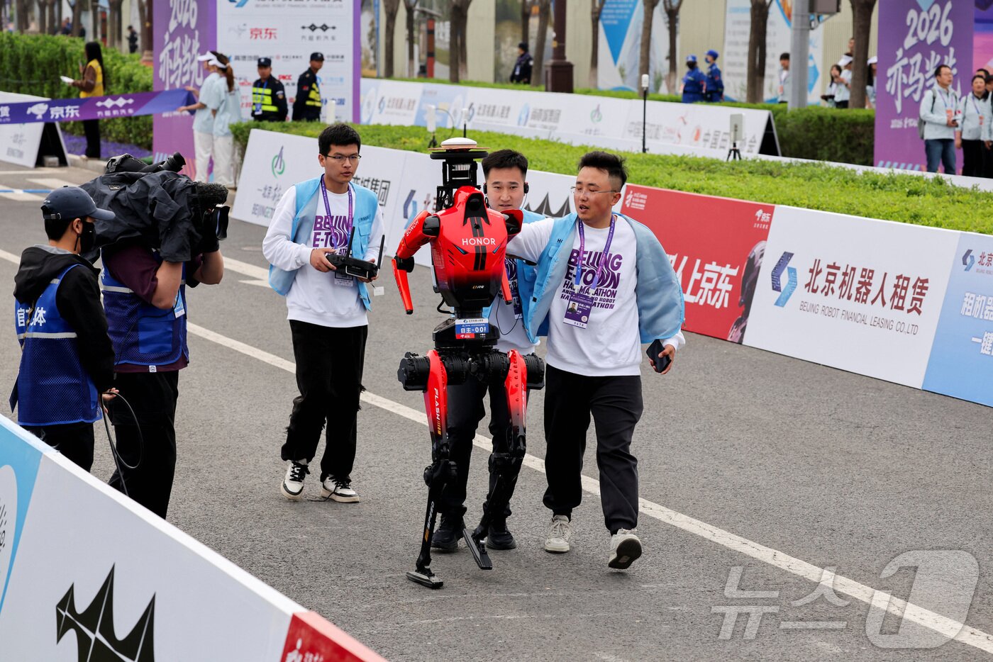 본문 이미지 - 19일 중국 베이징에서 열린 제2회 베이징 이타운 하프 마라톤 및 휴머노이드 로봇 하프 마라톤에서 우승한 아너 라이트닝 휴머노이드 로봇이 팀원과 함께 걷고있다. ⓒ 로이터=뉴스1
