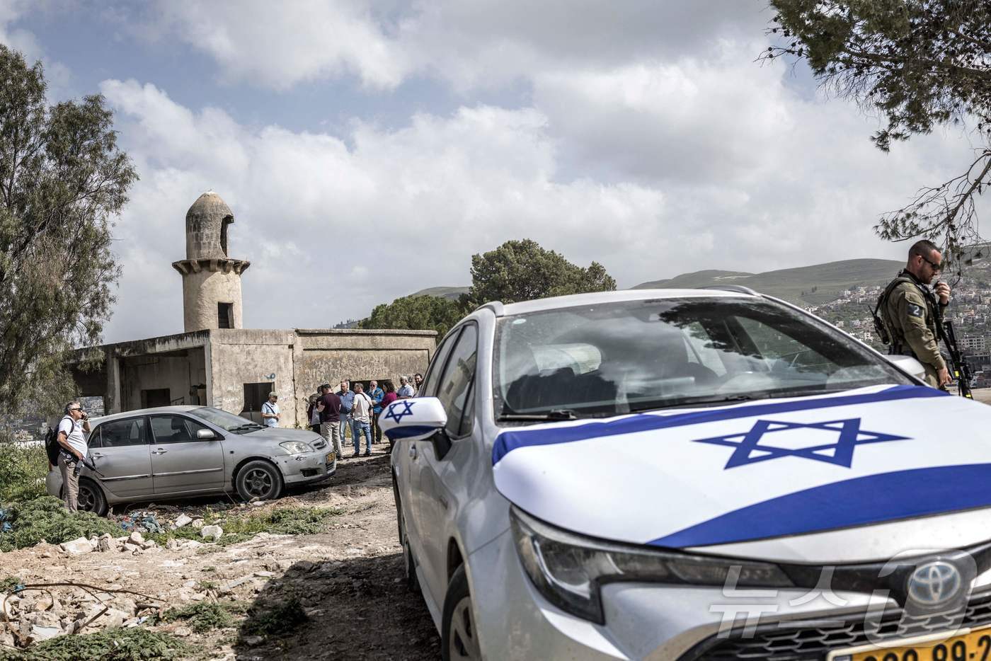 본문 이미지 - 19일(현지시간) 팔레스타인 서안지구 내 유대인 정착촌 사누르 재정착 기념식이 끝날 무렵, 한 이스라엘 군인이 이스라엘 국기로 덮인 차량 근처에서 무전기로 대화하고 있다. 그 뒤로 이스라엘 정착민 무리가 모스크 구조물 앞에 모여 있다. 2026.04.19. ⓒ AFP=뉴스1