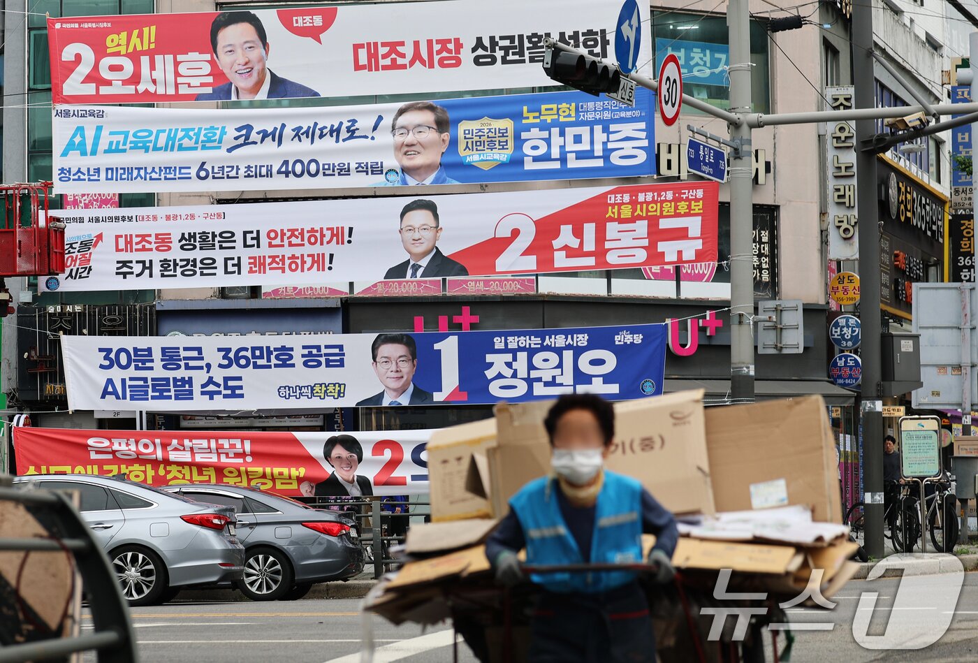 본문 이미지 - 6·3 지방선거 공식 선거운동이 시작된 21일 서울 은평구의 한 거리에서 폐지 수레를 끄는 할머니가 여야 후보자들의 선거 현수막 앞을 힘겹게 지나고 있다. 2026.5.21 ⓒ 뉴스1 오대일 기자