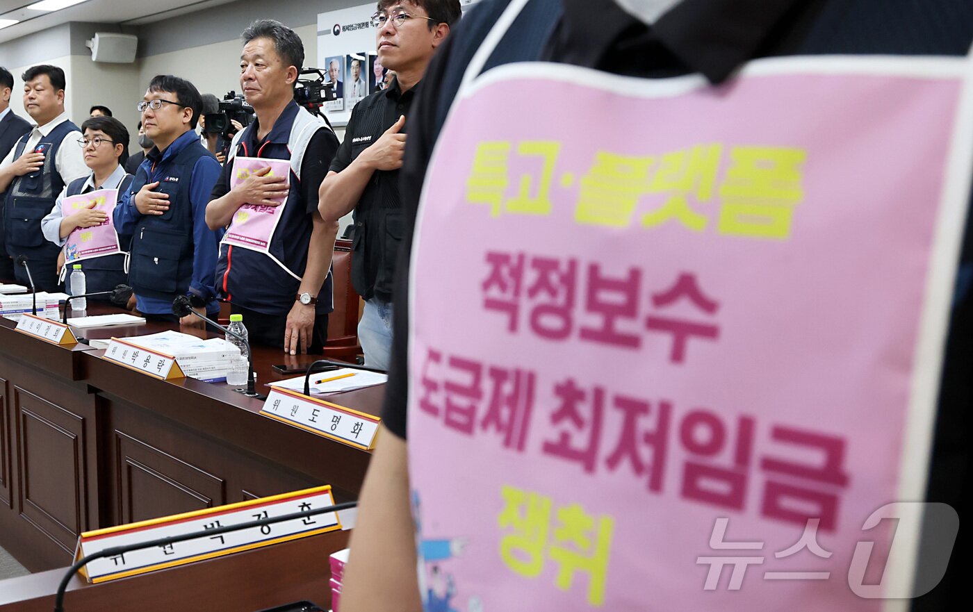본문 이미지 - 박정훈 근로자위원이 26일 세종시 정부세종청사 최저임금위원회에서 열린 2027년도 최저임금위원회 3차 전원회의에 참석하여 특고·플랫폼 적정보수 밒 도급제 최저임금 쟁취 문구를 착용한 채 국민의례를 하고 있다. 2026.6.4 ⓒ 뉴스1 김기남 기자